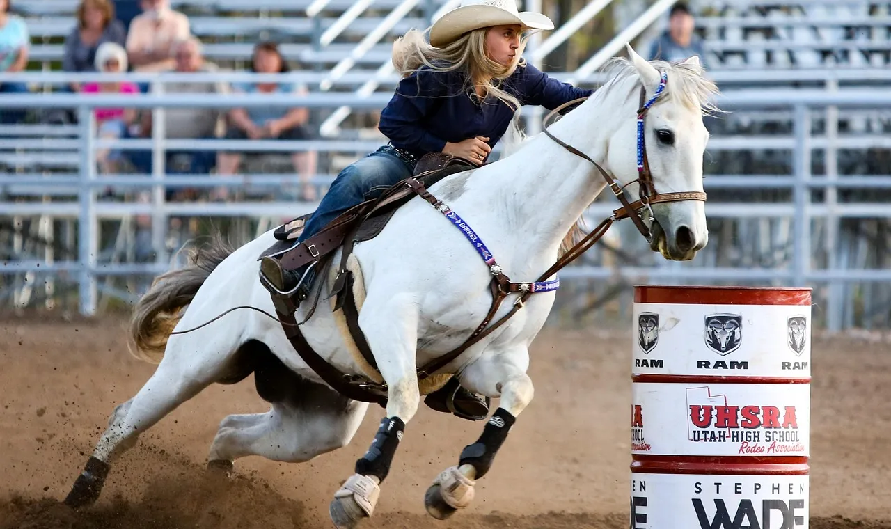 A young rider in a white hat on a grey horse leaning hard into a turn around a red and white barrel, dust kicking up behind