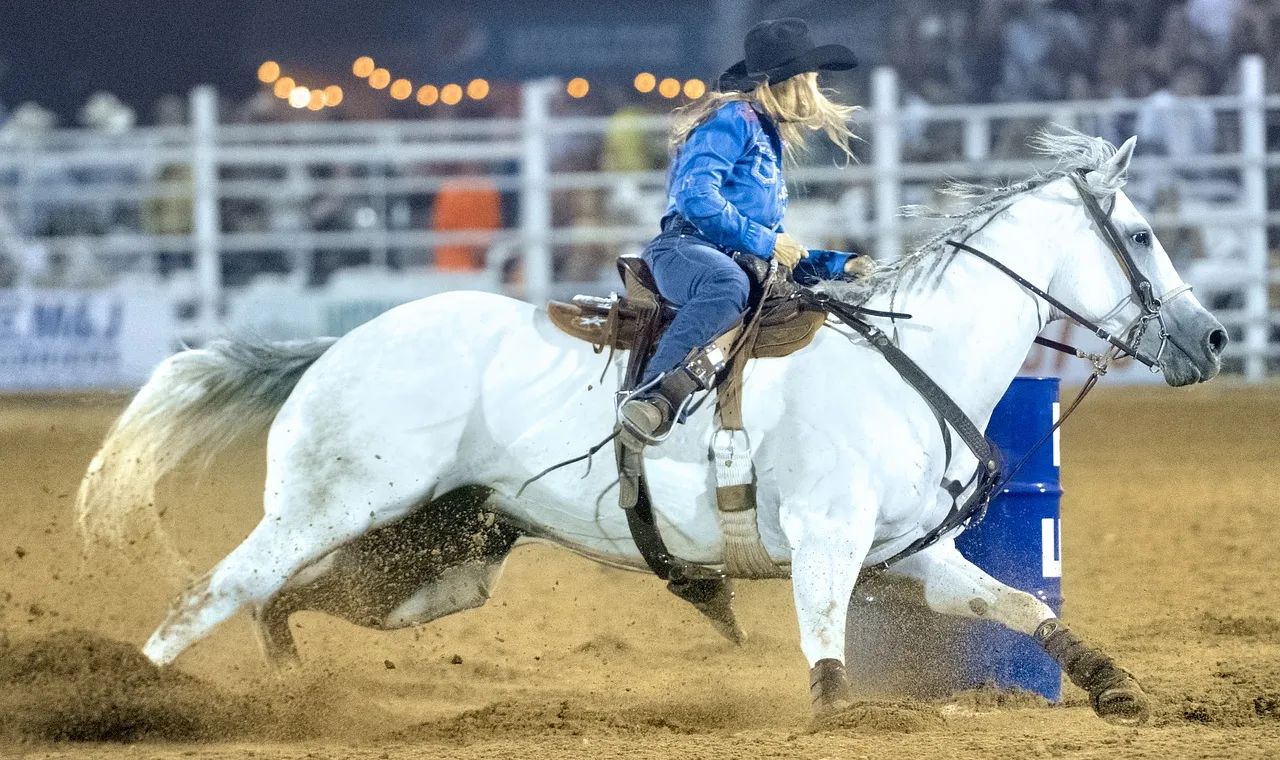 A rider on a grey horse turning tight around a blue barrel under arena floodlights, sand spraying from the horse's feet