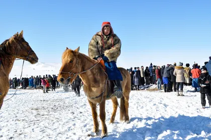 A young rider in a thick fur coat and red hat on a small stocky chestnut horse, standing on trodden snow in front of a crowd of spectators