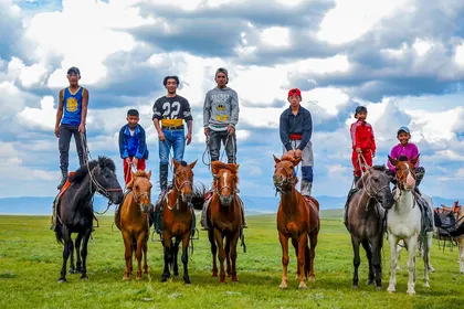 Seven Mongolian boys and young men on small horses on green summer steppe, several of them standing upright on the horses' backs