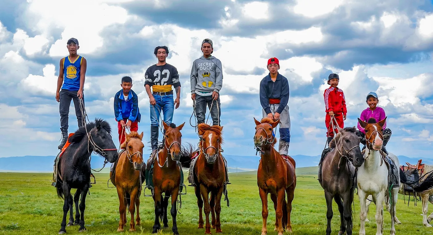 Seven Mongolian boys and young men on small horses on green summer steppe, several of them standing upright on the horses' backs